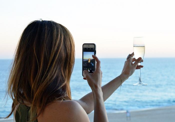 Bilder einer Frau, die mit Ihrem Handy ein Foto eines Sektglases am Strand macht