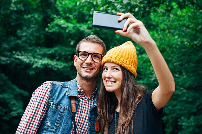 Bild eines lächelndes Paares, das vor einigen Bäumen ein Selfie macht