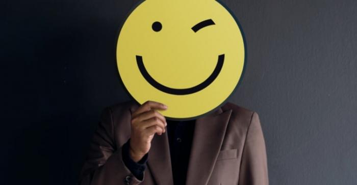 Bild eines männlichen Körpers in Anzug, der sich einen großen gelben zwinkernden Smiley vor das Gesicht hält